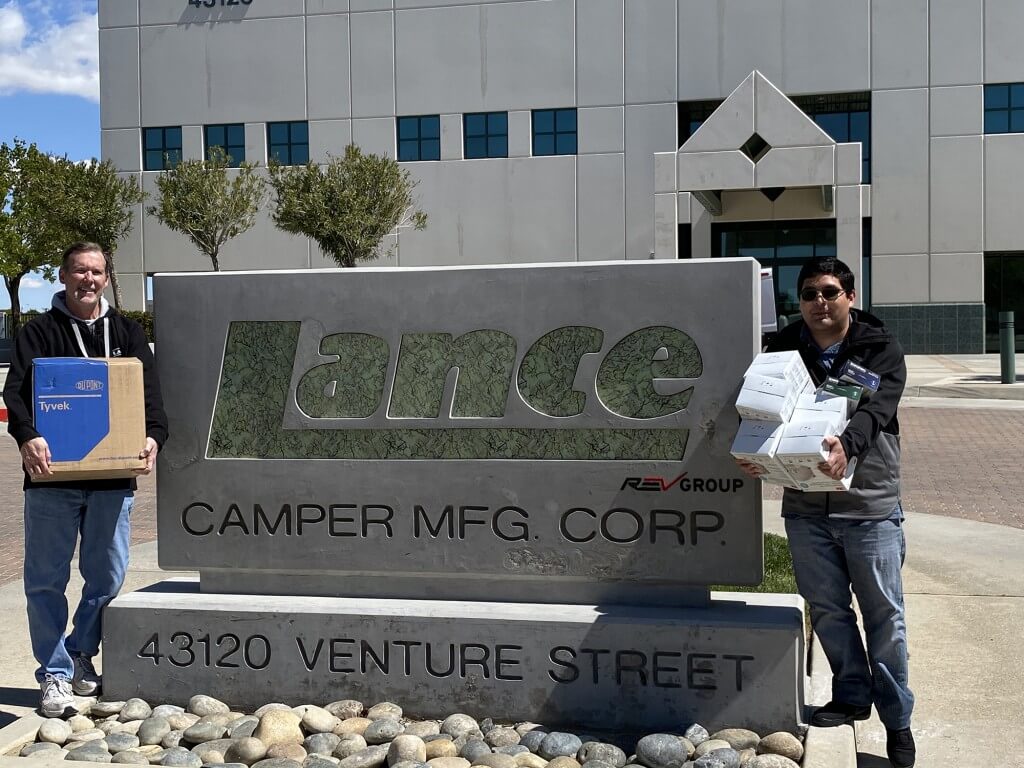 Two people standing in front of Lance sign by Lance Manufacturing plant holding hospital supplies that are being donated