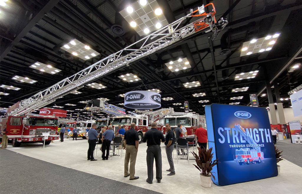 Image of people congregating around a E-ONE Fire Truck with its' ladder extended inside the conference center at FDIC 2022. E-One Tradeshow sign is in front of the fire truck.